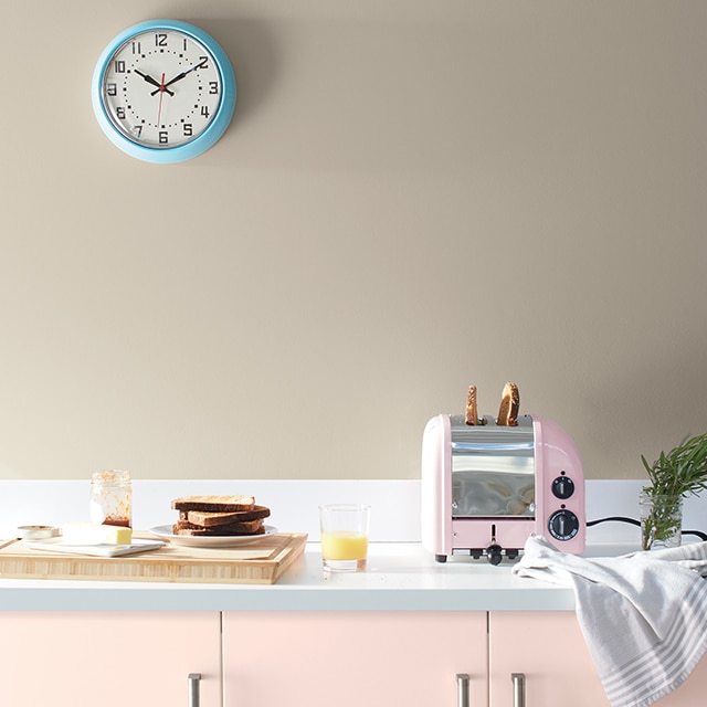 Une cuisine arborant des armoires peintes en rose pâle, un grille-pain rose, une planche à découper, de la vaisselle et un mur peint en brun clair avec une horloge bleue.