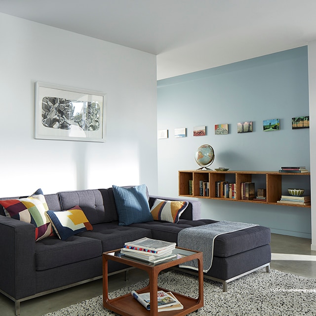Le soleil qui se reflète dans ce salon bleu pâle et blanc avec sofa modulaire gris foncé et coussins colorés éclaire aussi le mur bleu pâle du couloir qui présente une bibliothèque murale et des petits tableaux.