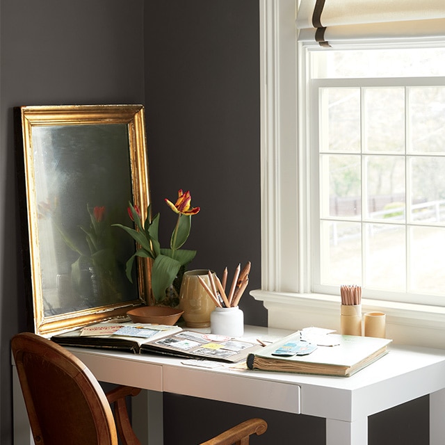 A pretty corner home office space with chocolate brown painted walls and a large, white-trimmed window in front of a white desk with scrapbooks and pencils, and a wood chair.