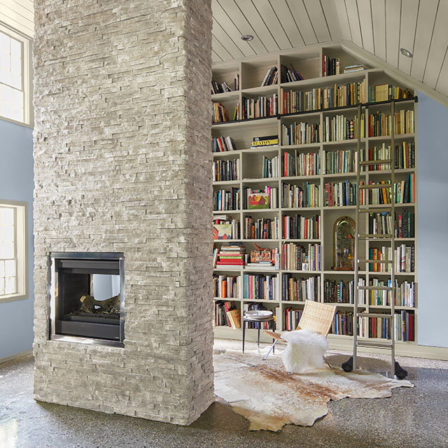 A home library with a statement floor-to-ceiling white-painted bookcase, blue walls, white shiplap vaulted ceiling and trim, and white stone center fireplace.