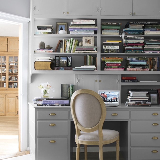 A home office area with built-in gray painted upper cabinets, shelves filled with books, a desk with drawers, a cushioned beige chair, and a pretty star-shaped chandelier.