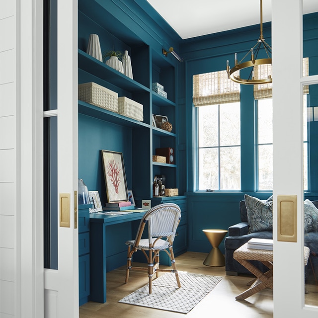 A beautiful teal-painted home office with built-in shelves, cabinets and desk, tall windows, beach décor, and open white pocket doors with brass hardware.