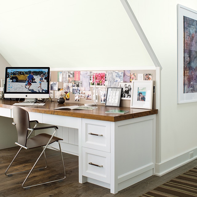 Bureau blanc avec dessus en bois, ordinateur et tableaux en dessous d’un mur incliné blanc et chaise brune en métal