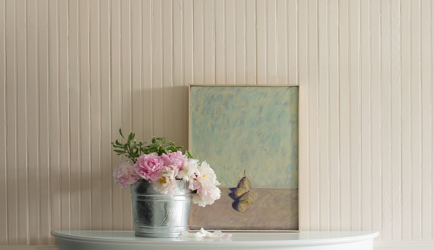 Pale pink walls with a painting and a tin bucket of pink flowers on top of a console.