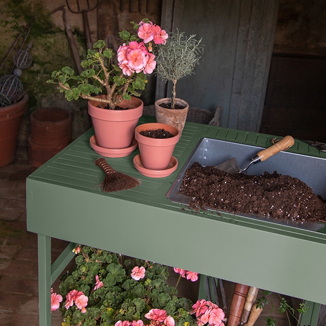 A green painted two-tiered plant stand with potted flowers and gardening tools.