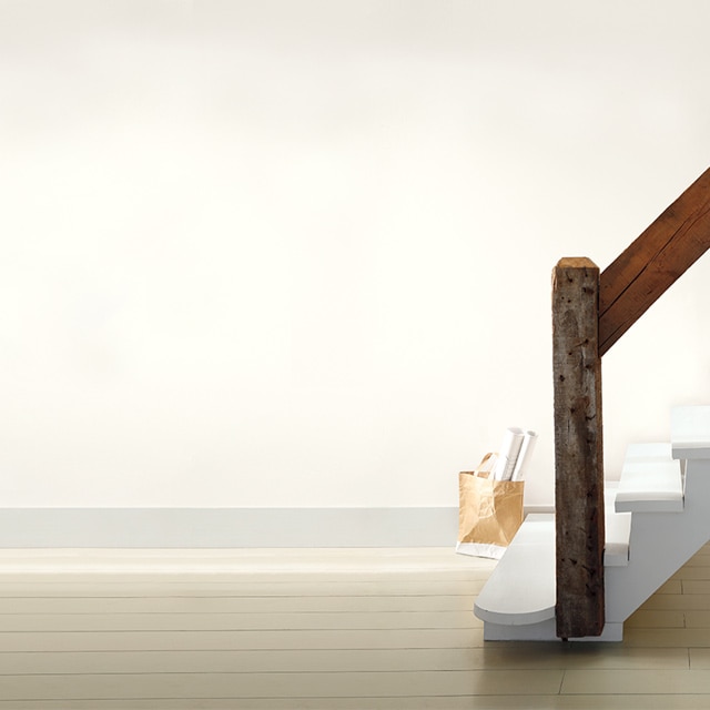 Un escalier blanc rustique muni d’une rampe en bois dans une pièce peinte en blanc cassé; un sac rempli d'affiches roulées est posé sur le parquet. 