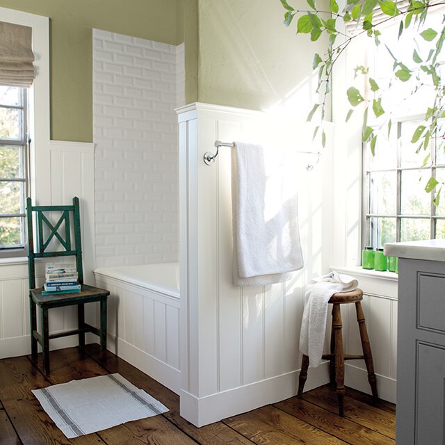 Salle de bains lumineuse aux murs beiges ornés de lambris blancs, qui présente une baignoire et une douche en alcôve, ainsi qu’une chaise et un tabouret en bois. 