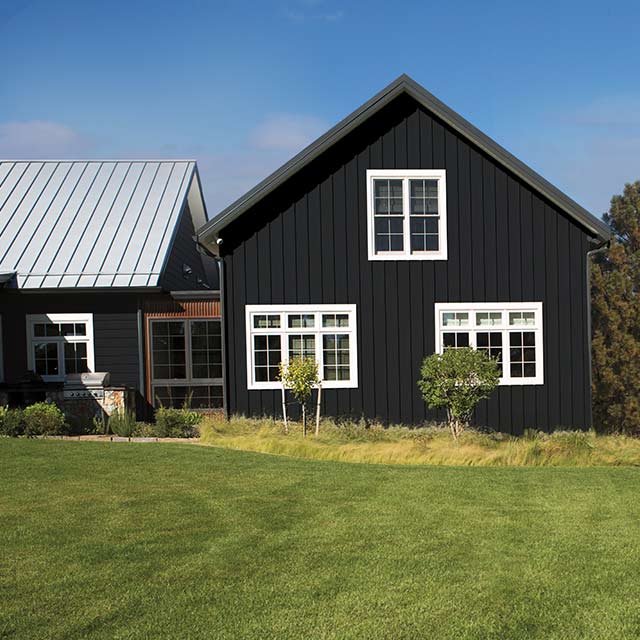 Une maison de ferme noire aux fenêtres blanches dans un champ, entourée de montagnes et de fleurs rouges par une journée ensoleillée.
