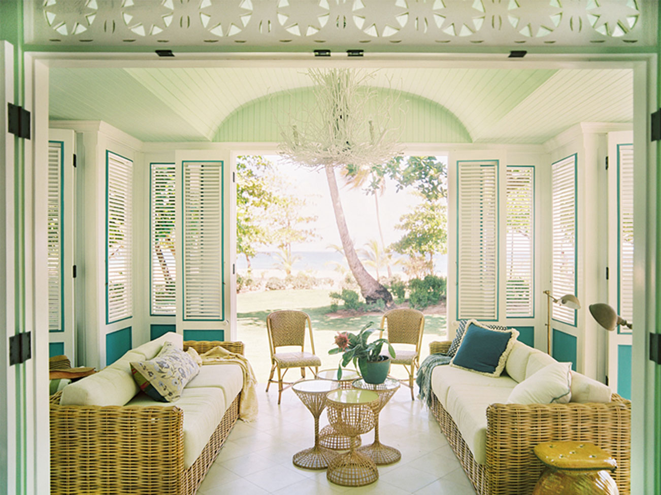 Véranda en forme d’îlot dans des teintes de vert et de blanc crème avec des volets à panneaux articulés, deux sofas en osier tressé et des chaises assorties.