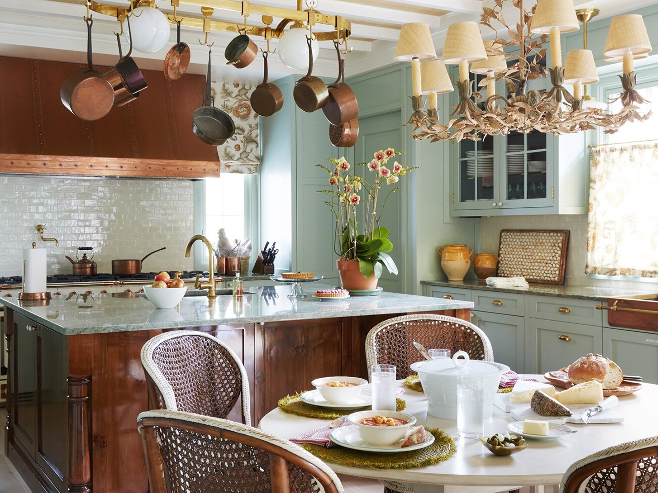 Cuisine au plafond à poutres apparentes avec hotte de cuisine en cuivre, grand îlot à base de bois et petite table élégante avec chaises de type bistro français.