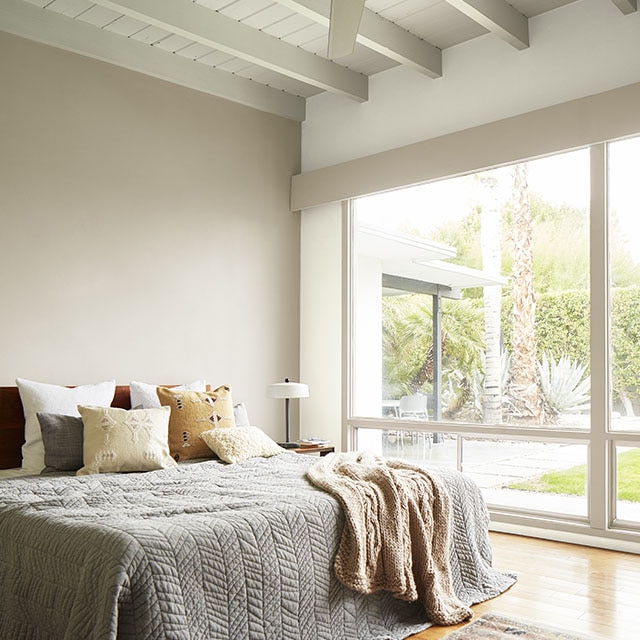 Chambre à coucher en teintes neutres et plafond en planches à feuillure blanches avec poutres exposées, ventilateur de plafond et fenêtre pleine longueur donnant sur un paysage désertique.