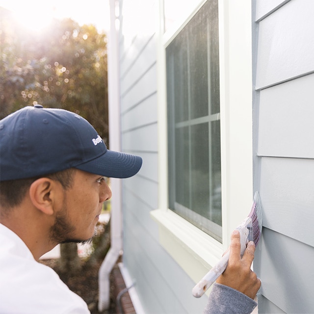 A professional contractor wearing a blue cap, applying light-gray paint to the exterior siding of a house near the edge of a white window.